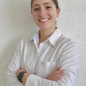 Portrait of Rebecca El Hawat wearing a white shirt, smiling at the camera with folded arms.