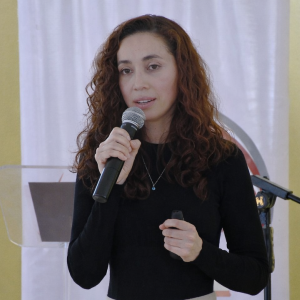 A woman speaking into a microphone during a presentation, holding a remote control while standing in front of a podium.