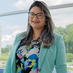 Portrait of a female scientist smiling indoors near a window, wearing glasses, a turquoise blazer, and a colorful blouse.