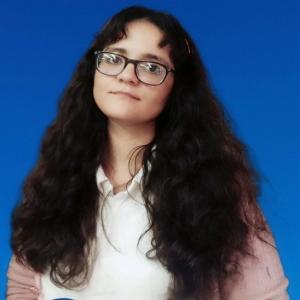 Portrait of Dr. Asma Raza with long dark wavy hair wearing glasses, a white shirt, and a light pink cardigan, standing against a solid blue background