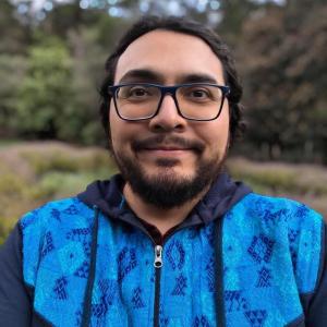 A cis man with glasess, black hair and barbe wearing a blue sweater. Behind him it is a forest.