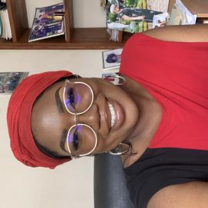 Professional headshot of Athina smiling, wearing glasses and a red headwrap with a red and black top, seated indoors with a bookshelf in the background.