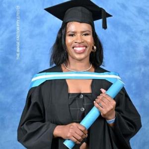 A smiling graduate in a black gown and cap, holding a blue graduation scroll with a light blue sash across the shoulders, standing against a blue background