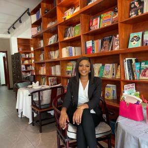 A woman in a black blazer and white blouse sitting on a wooden chair in a cozy bookstore or library, with tall wooden bookshelves filled with books behind her and a pink handbag placed on a nearby table.