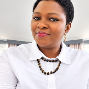 A professional head-and-shoulders portrait of a smiling woman with short natural hair, wearing a white collared shirt, pearl drop earrings, and a black-and-gold beaded necklace against a softly blurred indoor background