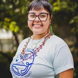 A Brazilian woman (with indigenous features) smiling at the camera, wearing glasses and a light-colored t-shirt with an ocean-related logo, standing outdoors with green vegetation in the background.