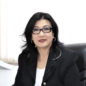 Photo of a professional woman with shoulder-length black hair and glasses, wearing a black blazer and white top, sitting in an office chair with a bright background.