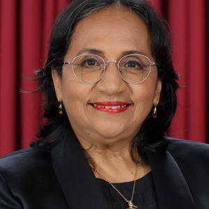 Ana Sandra de edad en vejez temprana, tiene una tez trigueña oscura, pelo ligeramente ondulado, longitud media, utiliza lentes y tiene una sonrisa agradable. su vestido es negro y el fondo de la foto es ocre. 