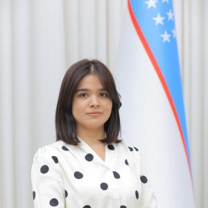Professional portrait of a woman wearing a white blouse with black polka dots, standing beside the flag of Uzbekistan against a light background.